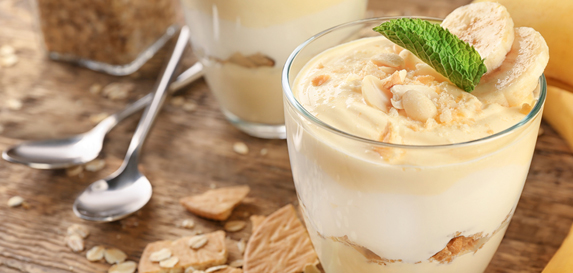Layers of banana pudding and whipped cream sit in glass cups on a wooden table with a banana.